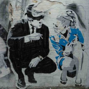 London Streetart Graffiti. Es zeigt das Bild eines Mannes und einer Frau, welche in der Hocke kniend miteinander sprechen. Der Mann trägt einen schwarzen Anzug und einen Hut und hält in seiner rechten Hand eine Zigarette. Die Frau trägt ein kurzes schickes blaues Kleid und hat eine Hochfrisur im Stil der 70’er Jahre. Im Hintergrund ist eine Graffiti Wand in den Farben, Schwarz-Weiß und Grau.