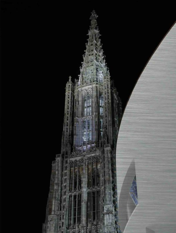 Ulmer Münster und Stadthaus Ulm. Es zeigt das Bild einer Nachtaufnahme des Ulmer Münsters mit seiner wunderschönen Lichtinstallation im Turm des Ulmer Münsters. Rechts im Bildteil ist das Ulmer Stadthaus, welches eine klare und moderne Architektur hat und komplett in Weiss ist. Entworfen wurde das Stadthaus vom New Yorker Architekten Richard Meier. Der Himmel im Hintergrund ist durchgehend schwarz.