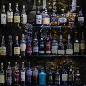 Edinburgh Bar Whiskyflaschen. Es zeigt das Bild von Whiskyflaschen, welche auf einem Regal vor einer dunklen Natursteinwand stehen. Es sind 3 Regalböden aus Glas, die alle gefüllt sind mit Whiskyflaschen vorrangig aus Schottland. Das Bild ist quadratisch und in Farbe.