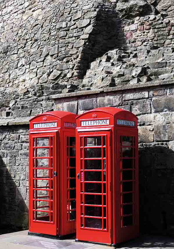 Schottland Edinburgh. Es zeigt das Bild von 2 britischen Telefonzellen, die nebeneinander vor einer grauen Natursteinwand, welche charakteristisch für Edinburgh ist, stehen. Die Telefonzellen sind in knalligem Rot. Das Bild ist im Hochformat und in Farbe.