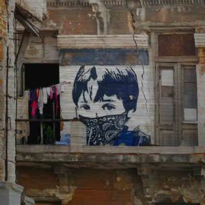 Havanna StreetArt. Es zeigt das Bild eines Graffiti’s an einem Haus in Havanna. Das Haus ist ziemlich verfallen, vom Balkon ist nur noch ein Teil vorhanden. Der Balkon ist noch begehbar, jedoch ist kein Geländer angebracht. Es ist eine Wäscheleine befestigt, an der verschiedene bunte Kleidungsstücke hängen. Das StreetArt mit dem Jungen ist in verschiedenen Blau- und Weißtönen. Es ist eine Porträtaufnahme des Jungen bis zu den Schultern zu sehen, er trägt eine Kurzhaarfrisur, jedoch trägt er ein Blau-Weißes Tuch über Nase und Mund, welches an einen maskierten Guerillakämpfer erinnert. Das Alu Dibond Bild ist quadratisch.