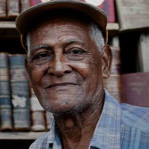 Havanna Model. Es zeigt die Porträtaufnahme eines alten Mannes aus Havanna, dessen Gesicht vom Leben gezeichnet ist. Er trägt eine Schildmütze und ein Blau/Weiß/Flieder gestreift/kariertes Herrenhemd, welches bis zur Schulter sichtbar ist. Die Hautfarbe des Mannes ist dunkel und im Hintergrund sind unscharf ältere Buchrücken zu sehen. Das Alu Dibond Bild ist im Querformat.