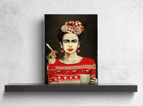 Berlin StreetArt Frida. Es zeigt das gemalte Bild einer Frau, welche die mexikanische Malerin Frida Kahlo in moderner Kleidung darstellen soll. Die Frau ist bis zur Taille abgebildet und hat in der rechten Hand eine Zigarette und in der linken Hand eine große Kaffeetasse. Sie trägt einen Pullover mit winterlichen Motiven in den Farben Rot, Weiß und Schwarz. Außerdem runde Ohrringe in den selben Farben. Die Haare sind schwarz und in einer Zopfkrone nach oben geflochten. Diese sind jedoch nur seitlich sichtbar, da direkt auf dem Kopf 7 verschiedene Kugeln in den Farben Weiß und Rot abgebildet sind. Der Hintergrund ist schwarz. Das Holzbild ist im Hochformat und steht und auf einem schwarzen Wandregal vor einem weißem Hintergrund.